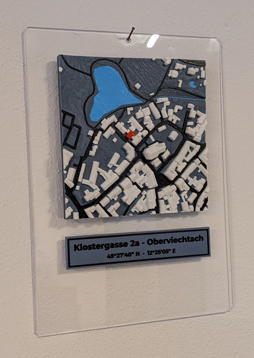 Relief einer Altstadt mit See und markiertem Haus auf einer klaren Acrylglasplatte an der Wand, darunter ein Schild mit Adresse und Koordinaten
