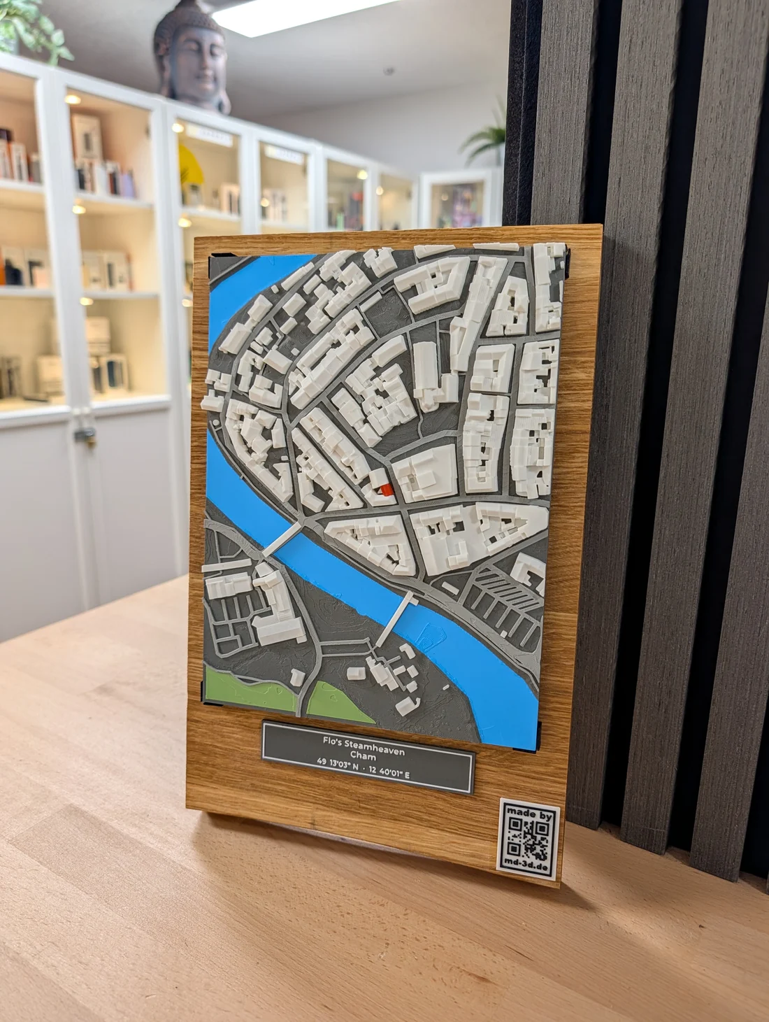 Farbiges 3D-Relief der Altstadt von Cham mit Fluss und markiertem Geschäft auf einem Eichenbrett, darunter ein Schild mit Name und Koordinaten, im Laden aufgestellt